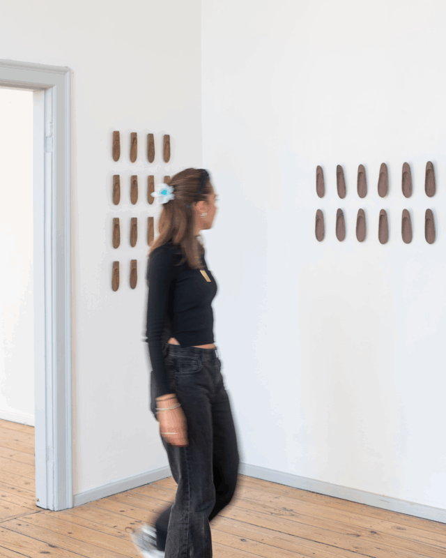 DH _ IG-2 - direktorenhaus A person in a black crop top and jeans walks past a wall displaying several brown art pieces in a gallery setting.