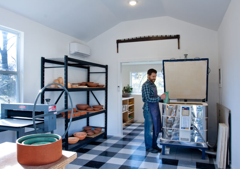 pintz_studio_portrait - direktorenhaus A man in a checkered shirt operates a kiln in a bright pottery studio with shelves of clay pots.